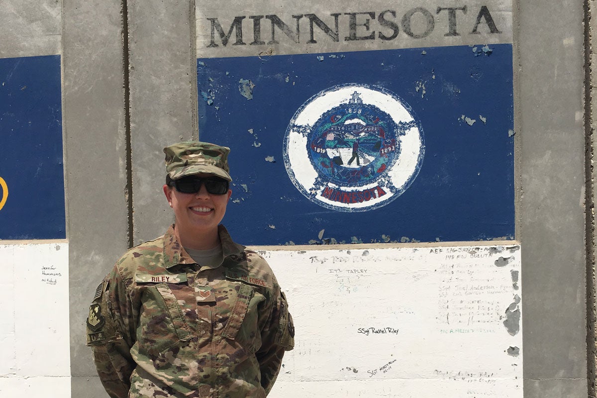 A woman in camouflage clothes and a hat and dark sunglasses stands in front of a concrete wall with Minnesota written on it