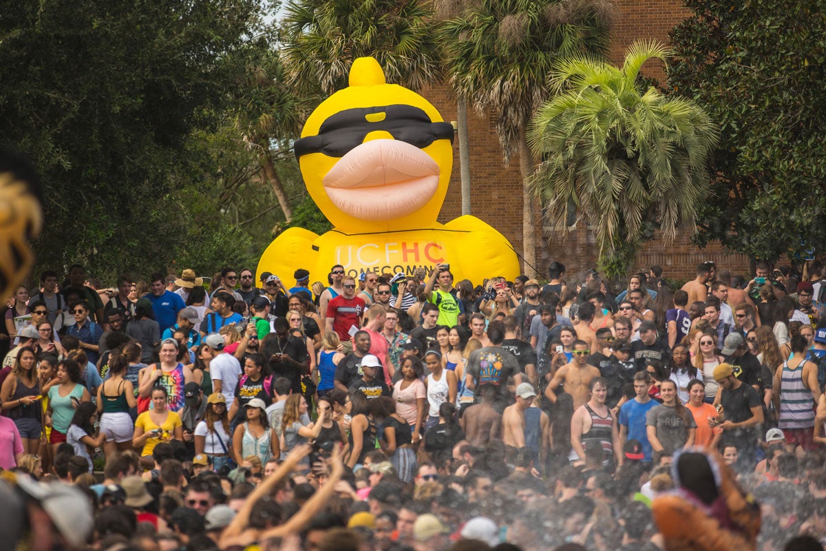 a giant inflatable yellow rubber duck wearing black sunglasses looms large over a crowd of college students