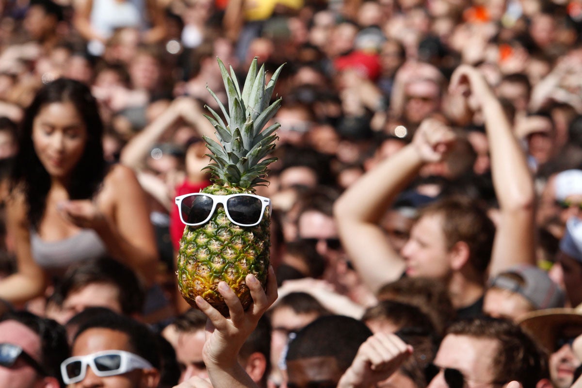 A hand holds up a pineapple wearing white sunglasses in a crowd of people