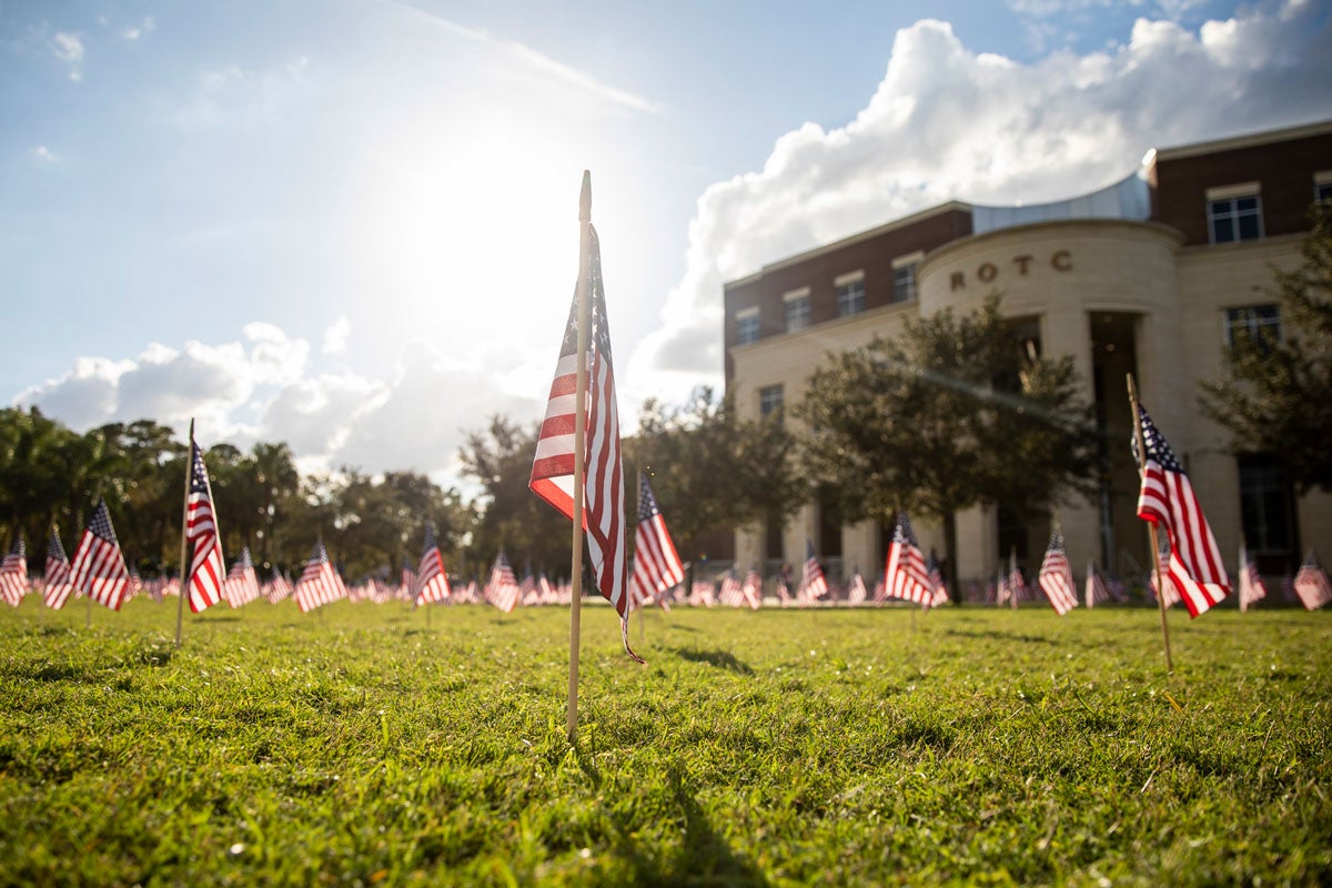 Sunny day, small American flags planted into grass with an ROTC brick building in the background