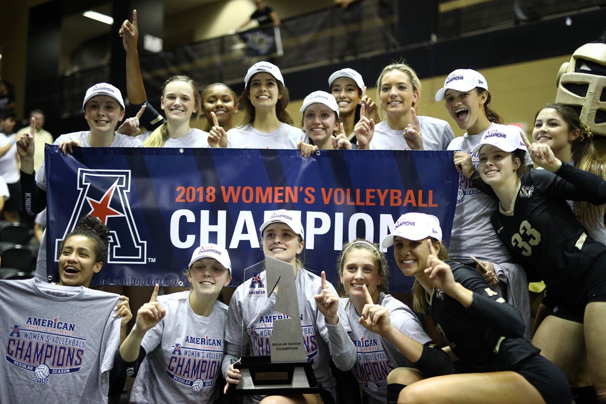 Volleyball team wearing white hats and gray t shirts pose with a silver trophy around a blue banner reading 2018 Women's Volleyball Champions 