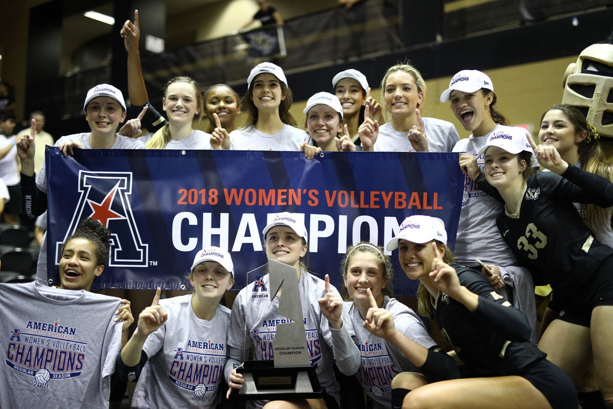 Volleyball team wearing white hats and gray t shirts pose with a silver trophy around a blue banner reading 2018 Women's Volleyball Champions