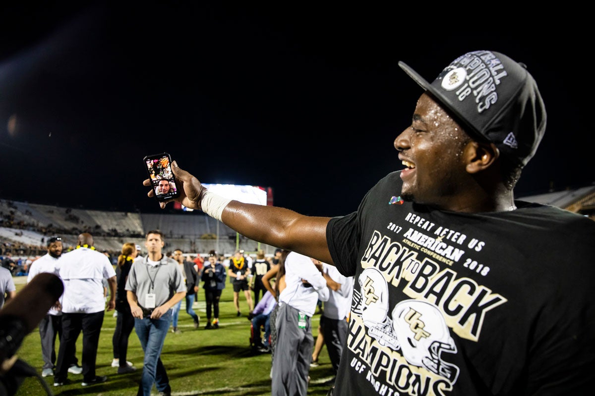 A UCF player wearing a black championship t shirt and hat holds out his arm to FaceTime McKenzie Milton