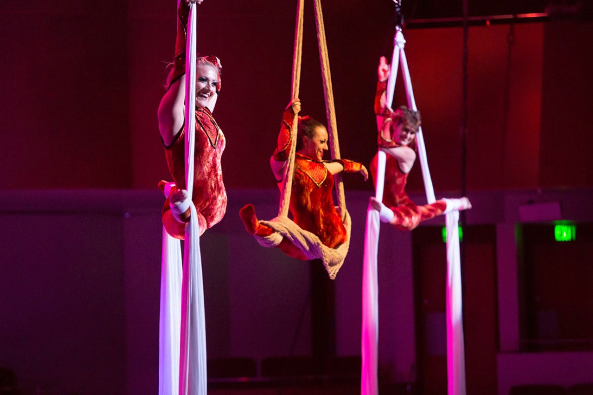 Three women wearing red costumes split midair while hanging from ropes