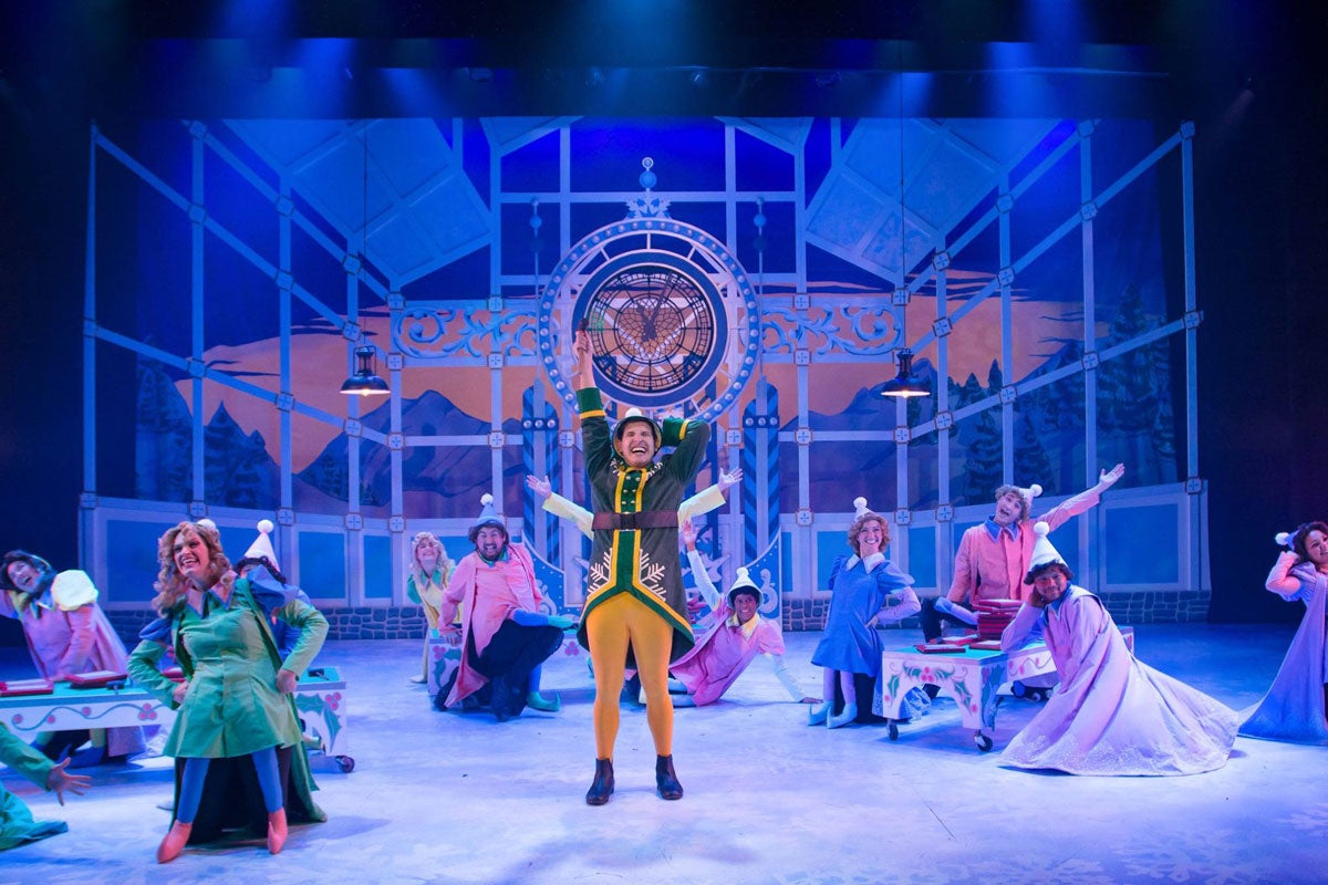 Theater stage with a man dressed like an elf in yellow pants and green jacket in the center standing and singing