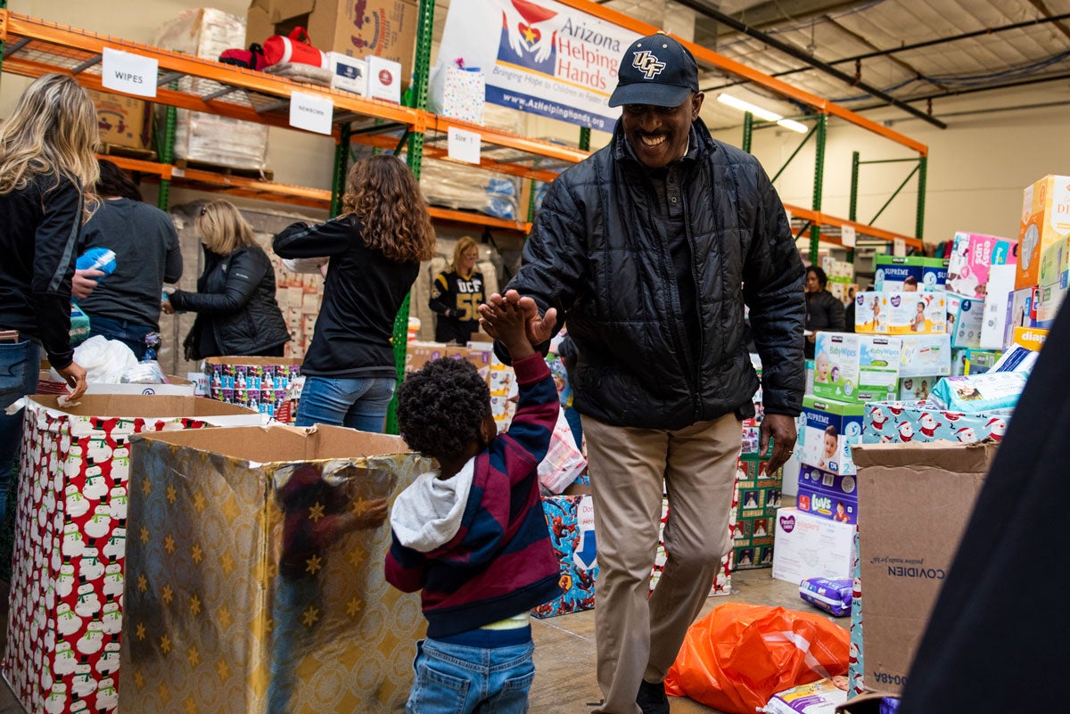 An adult male wearing a UCF hat and black shirt high fives a young boy in a warehouse.