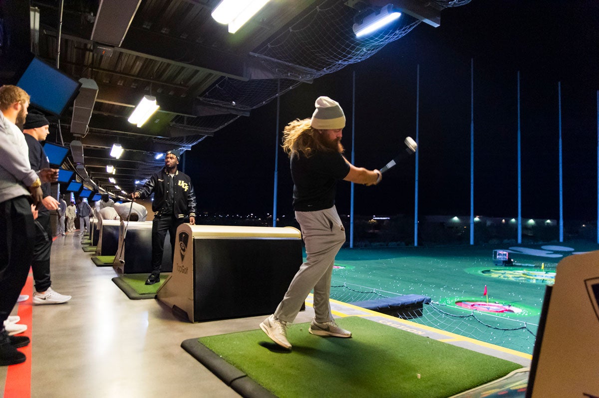 A man with long hair wearing a beanie and black shirt and khaki pants takes a golf swing at a driving range at night
