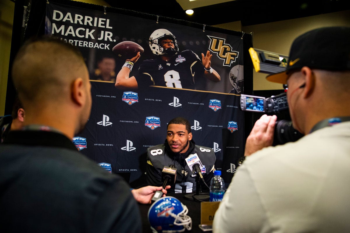 A football player sits in front of a media backdrop and answers questions into microphones from media members