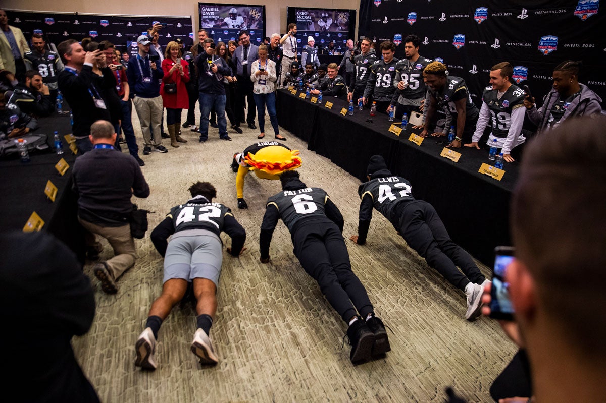 Three football players and a yellow sun mascot do pushups in front of a room of spectators