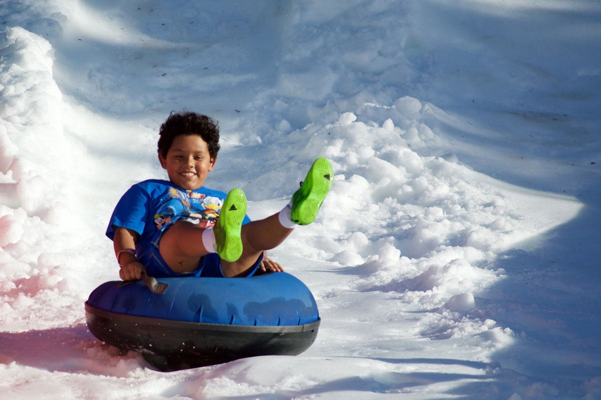 A young boy wearing a blue shirt and green shoes rides a snow tube