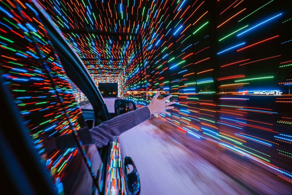 A hand and arm sticks out of a passenger window in a car with blurred colorful lights that make a tunnel