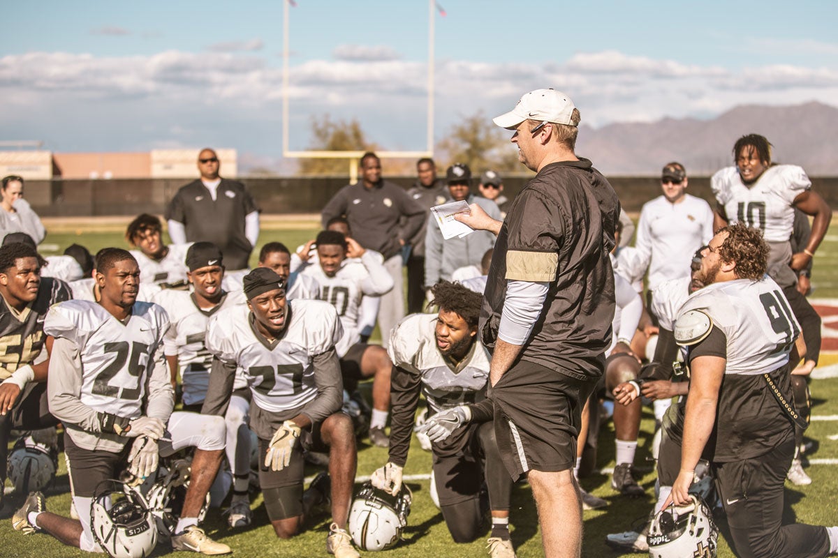 UCF head coach Josh Heupel, wearing white hat and black shirt and shorts, addresses players kneeling on field.