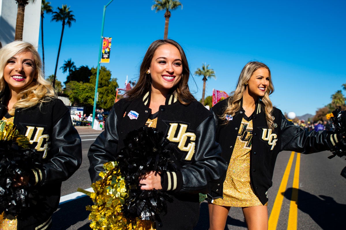 Three cheerleaders wearing black letterman jackets walk down a parade route.