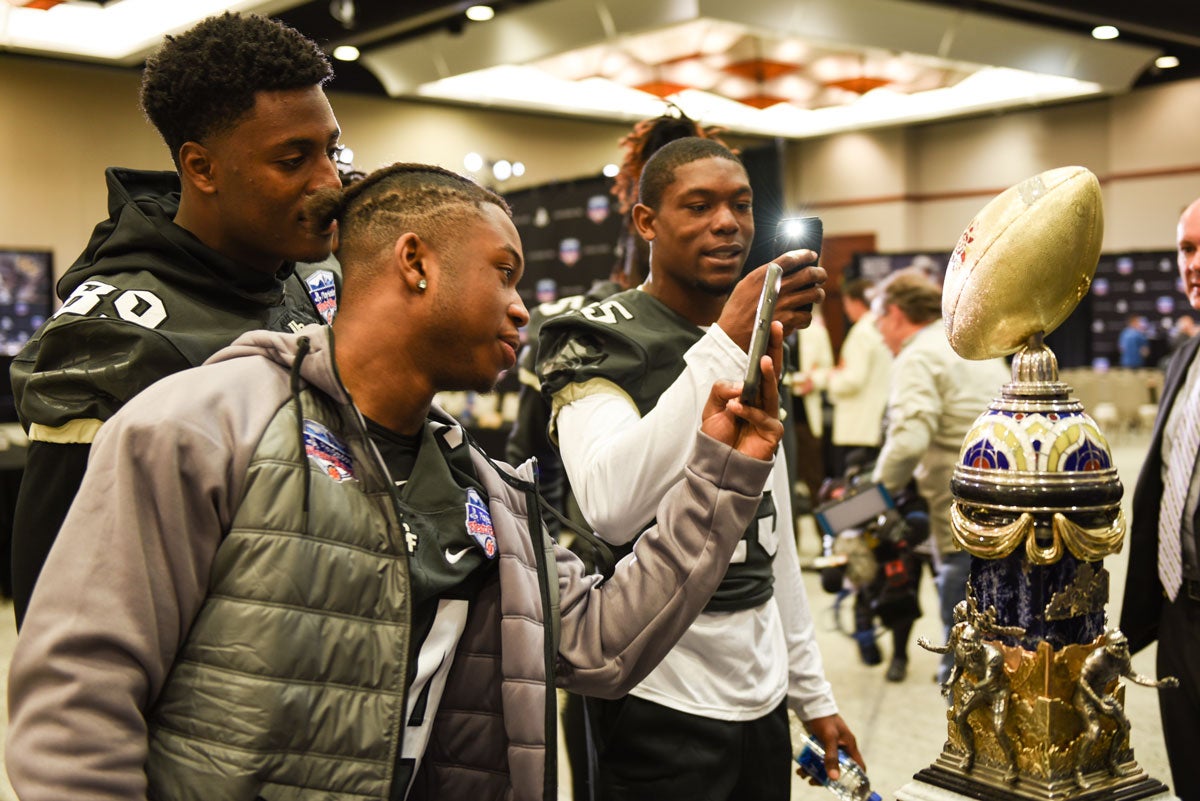 Three football players take photos with their cell phones of gold football-shaped Fiesta Bowl trophy