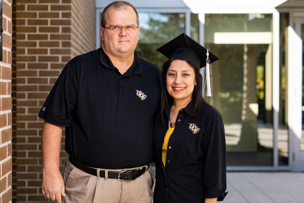 A tall man wearing a black t shirt, glasses and khaki pants puts his arm around a woman wearing a black graduation cap and black long sleeve shirt in front of a brick and glass building