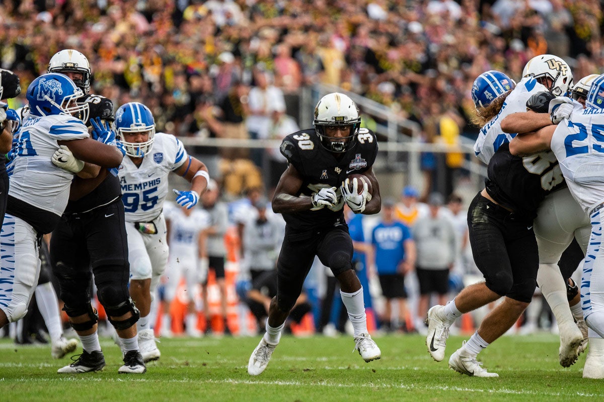 Running back Greg McCrae wearing a white helmet and black uniform runs through a hole one the field as his teammates block Memphis defenders on either side of him.