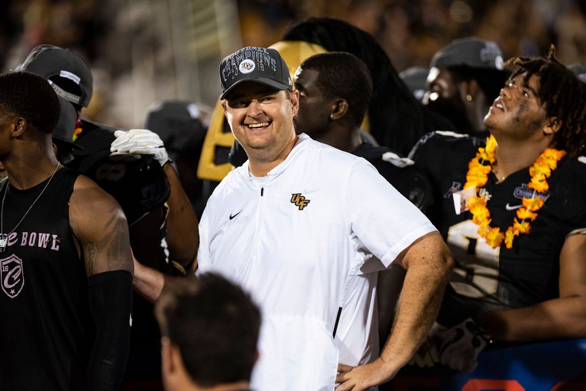 football coach Josh Heupel wearing a white polo shirt and gray hat poses with hand on hip and smiles