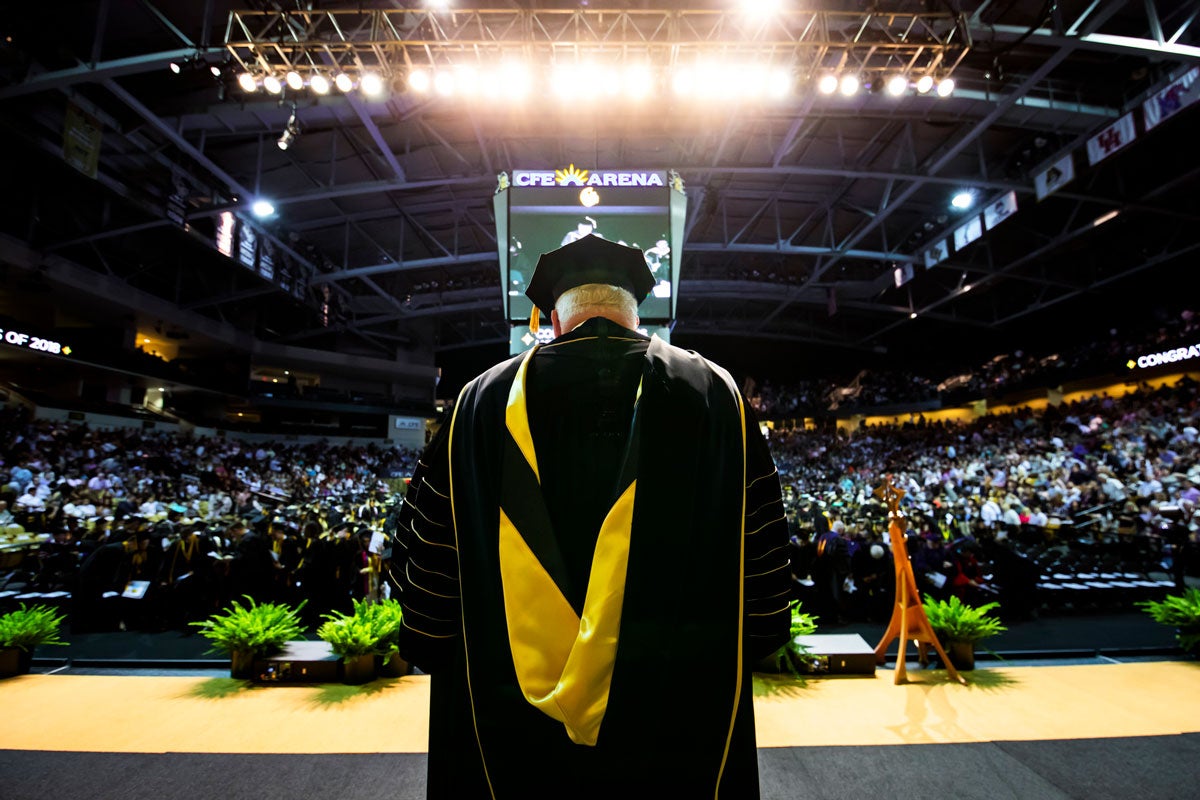 A man wearing graduation regalia stands on a stage and looks out at a crowded arena