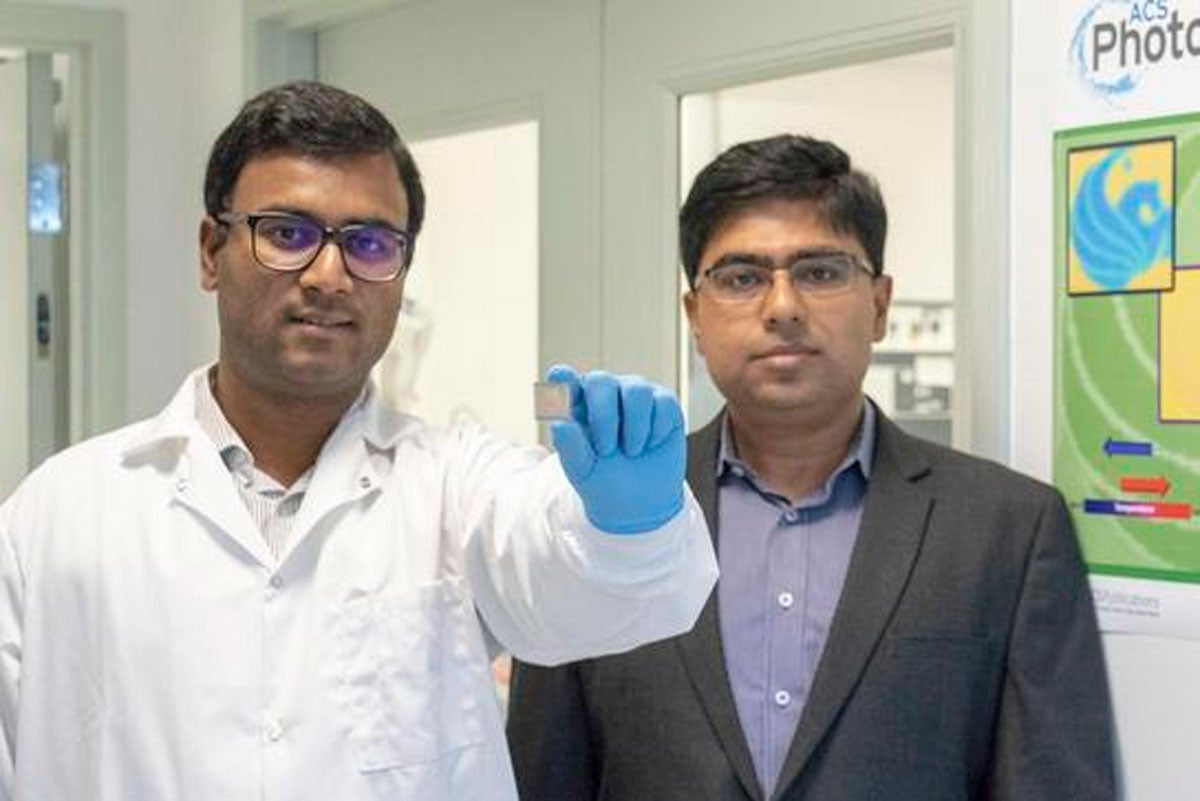 Two men stand side by side, the one on the left wears a lab coat and blue gloves and holds a small computer chip toward the camera