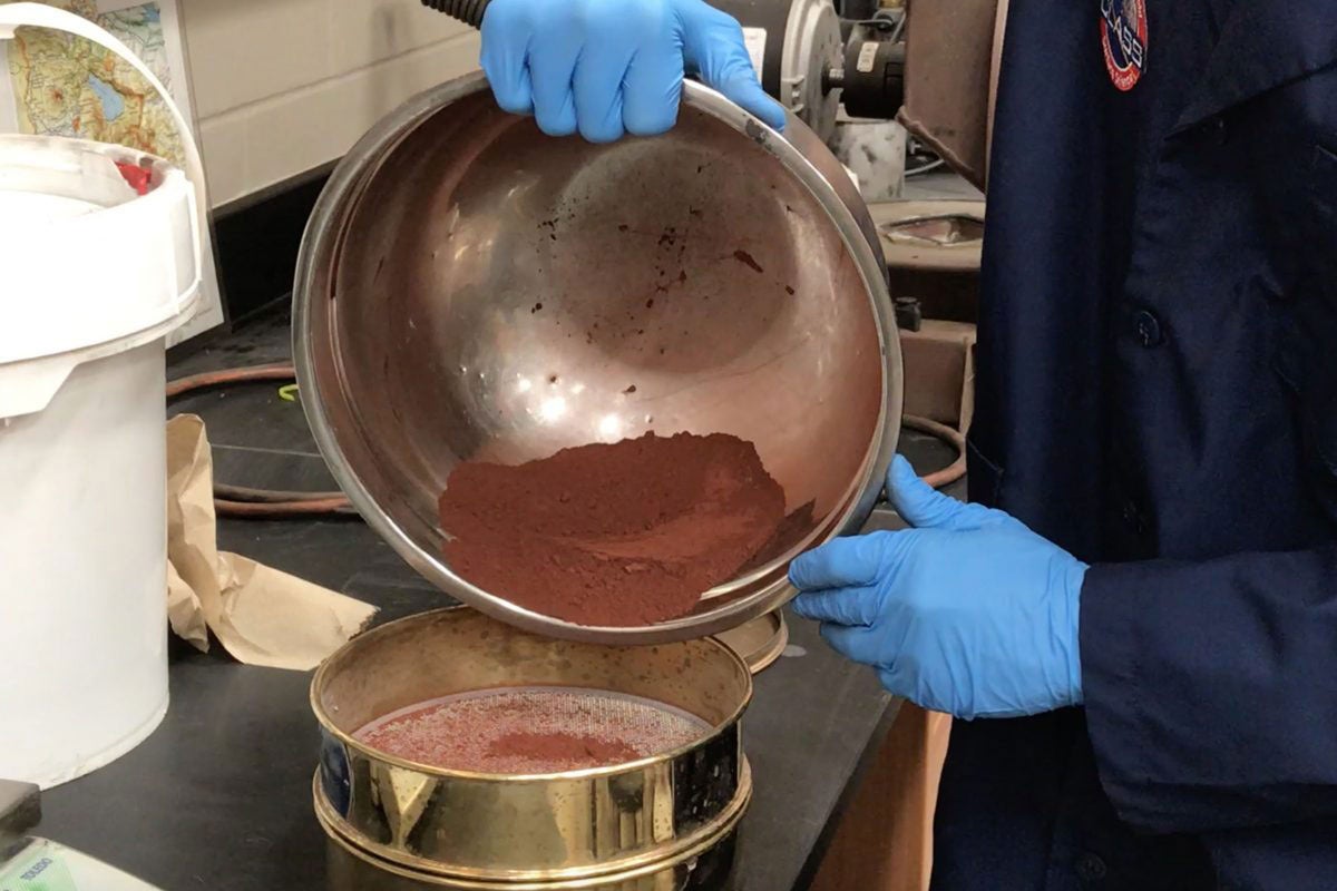 Hands wearing blue gloves hold a metal bowl filled with red-brown dirt