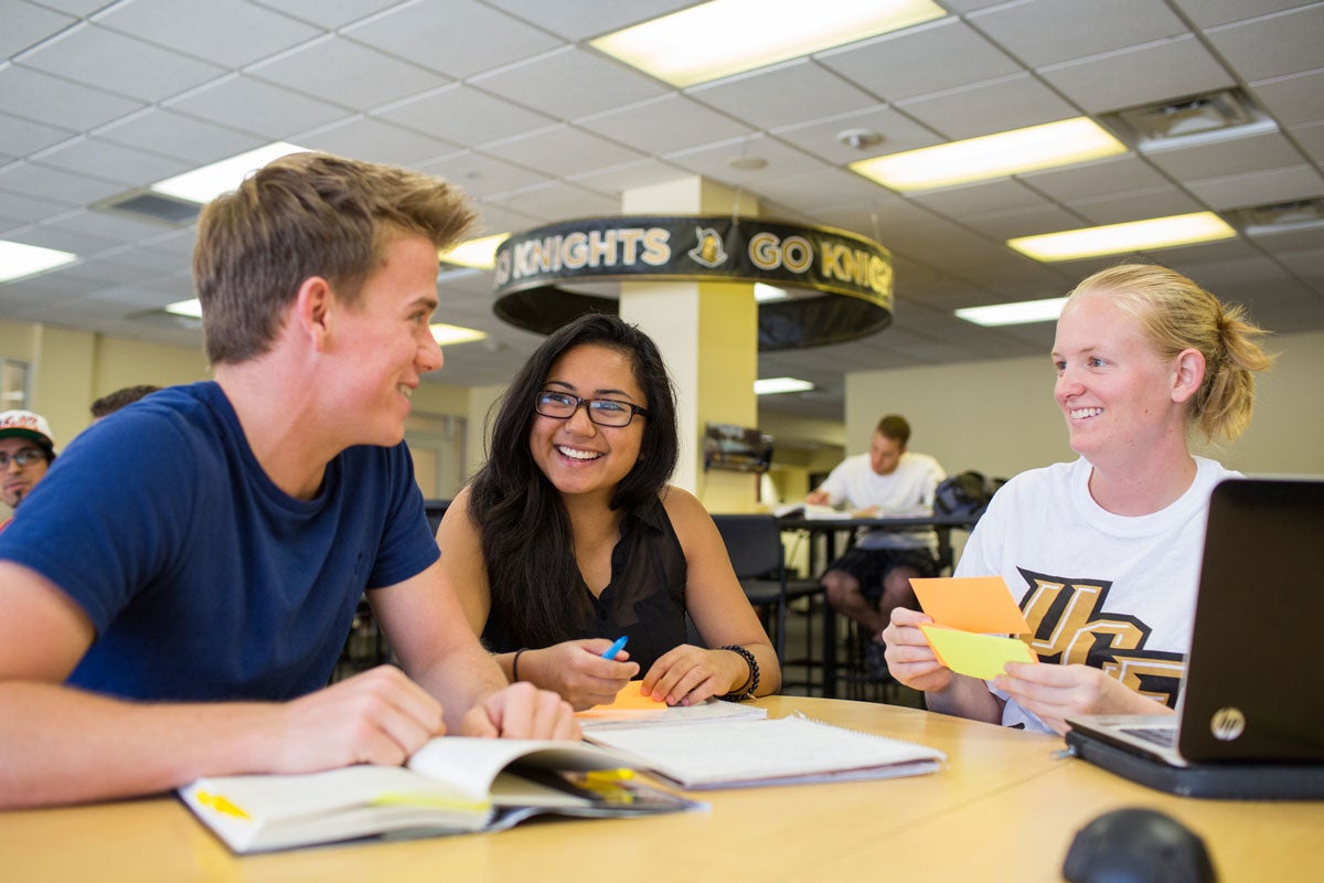 three students sit at a wood table with a laptop, books and flash cards