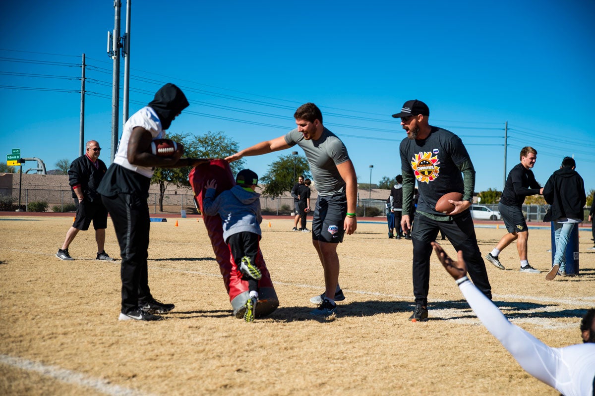 A child tackles a tackling dummy with football players supervising on a dirt lot