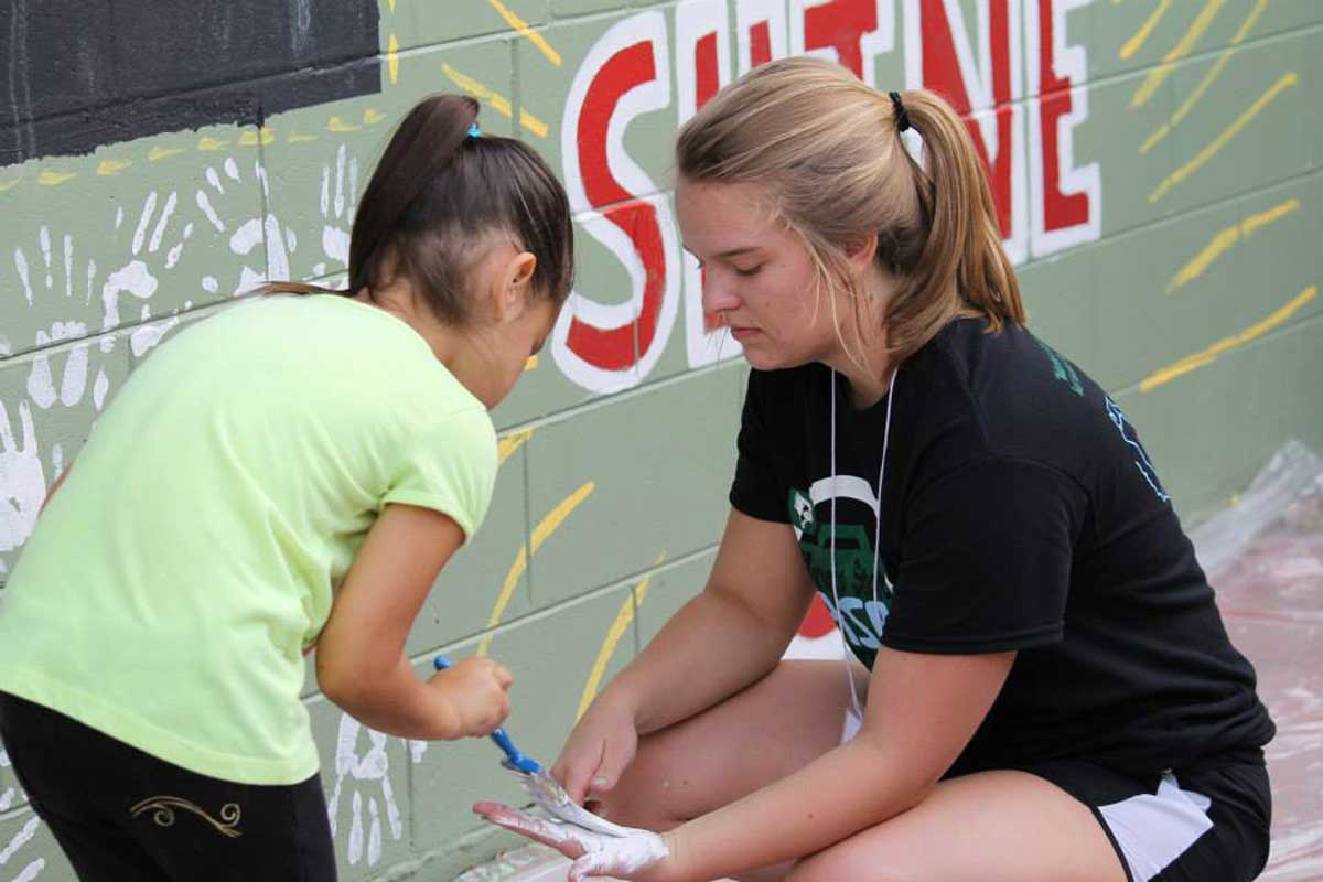 A young girl wearing a green shirt paints white paint with a blue brush onto the hand of a college-aged girl in front of a green wall with the word "shine" painted in red letters.