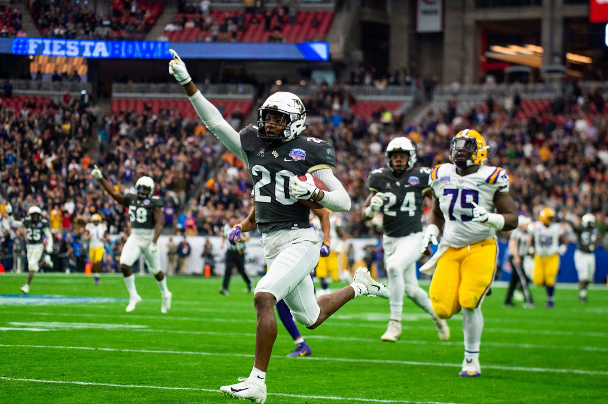 UCF football player wearing #20 black jersey raises right hand in the air in triumph as he runs into end zone with LSU players trailing