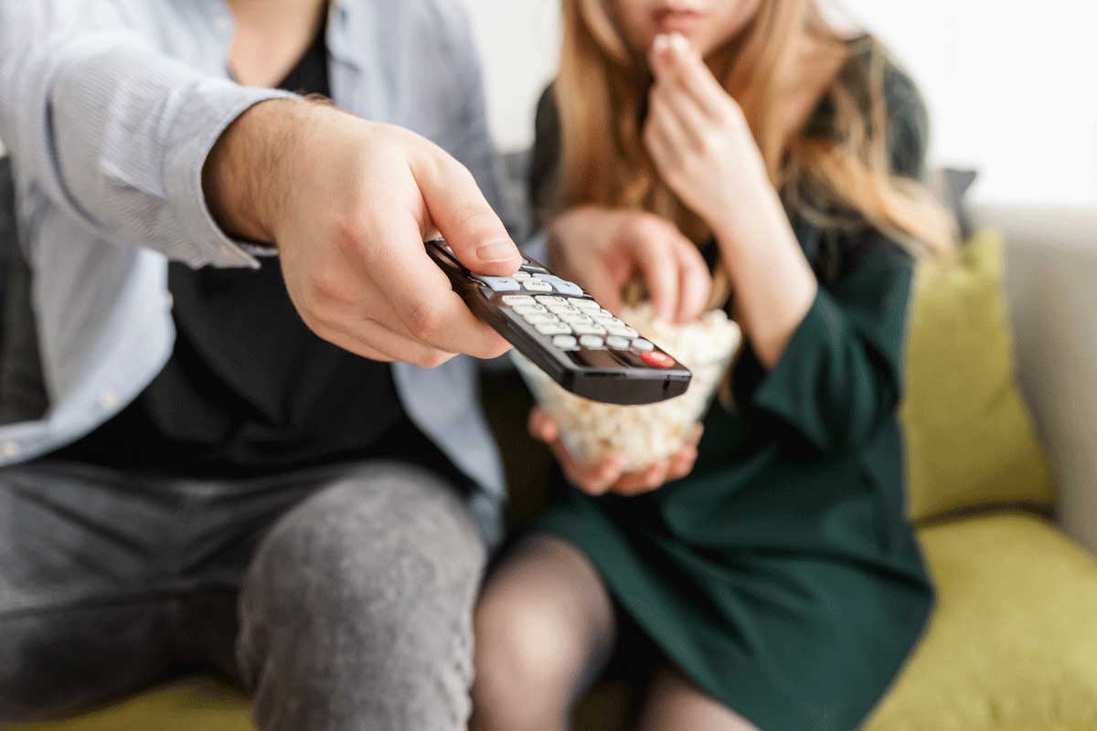two people sit on a couch and point remote control