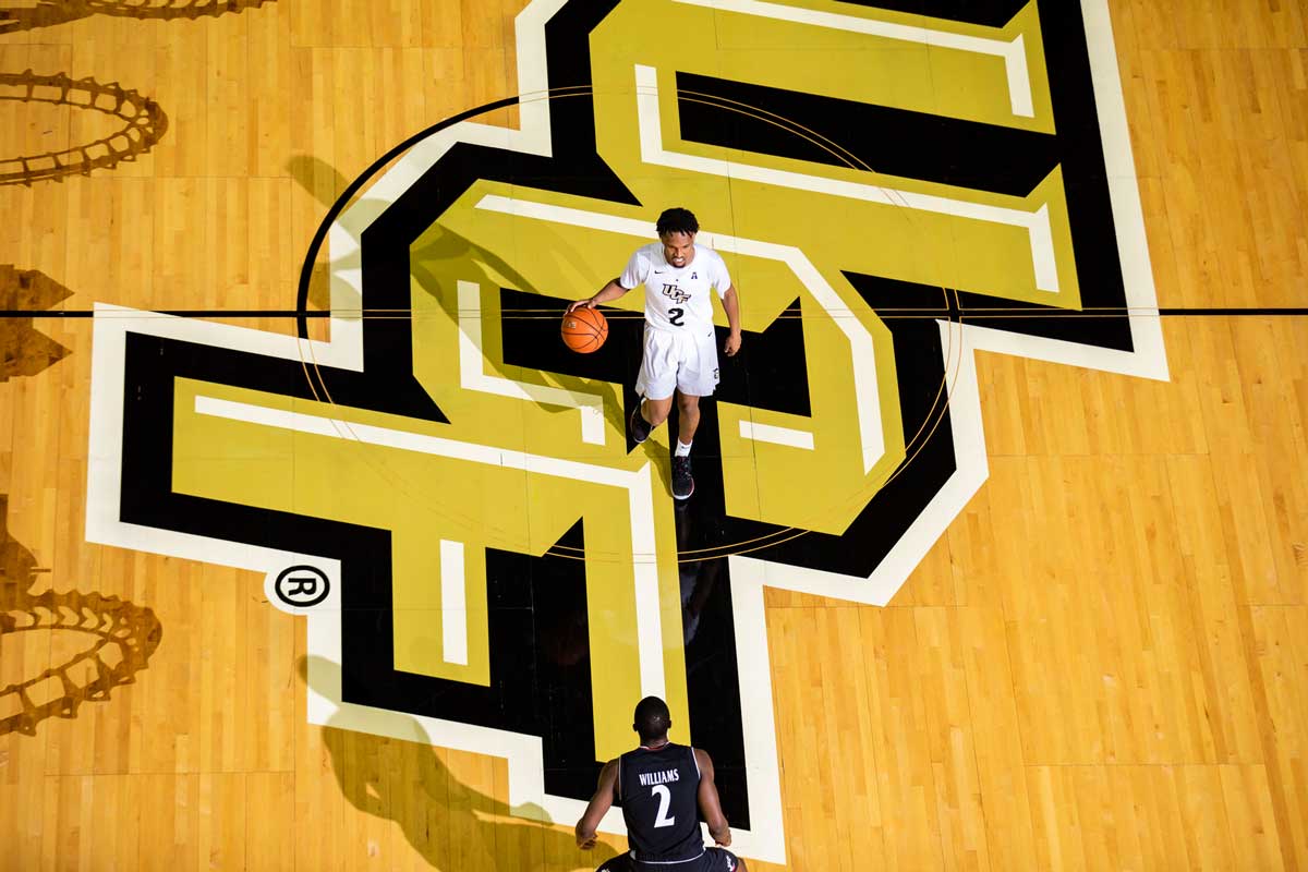 basketball player wearing white uniform dribbles ball across half court with UCF logo on floor