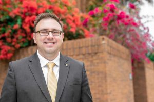 Man wearing glasses and gray suit with yellow tie stands in front of brick flower bed