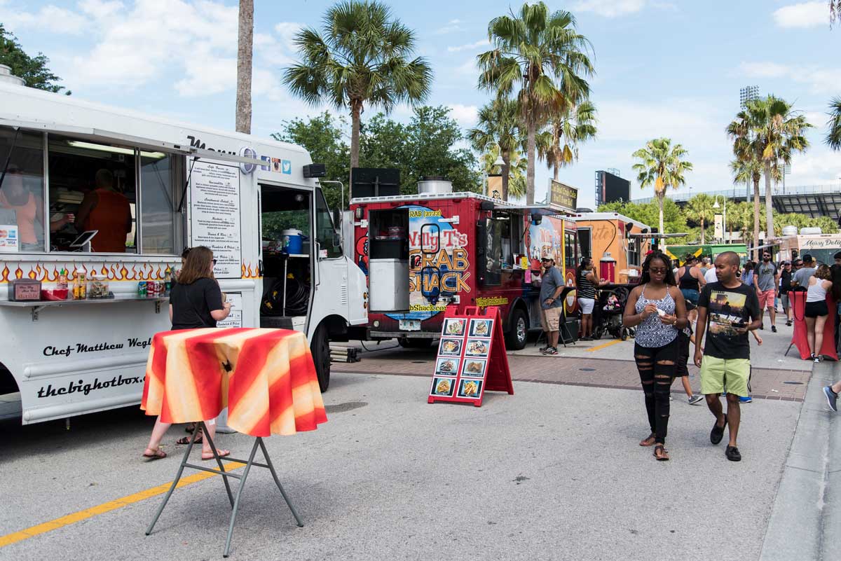 Food trucks lined up on a street with palm trees in background on sunny day