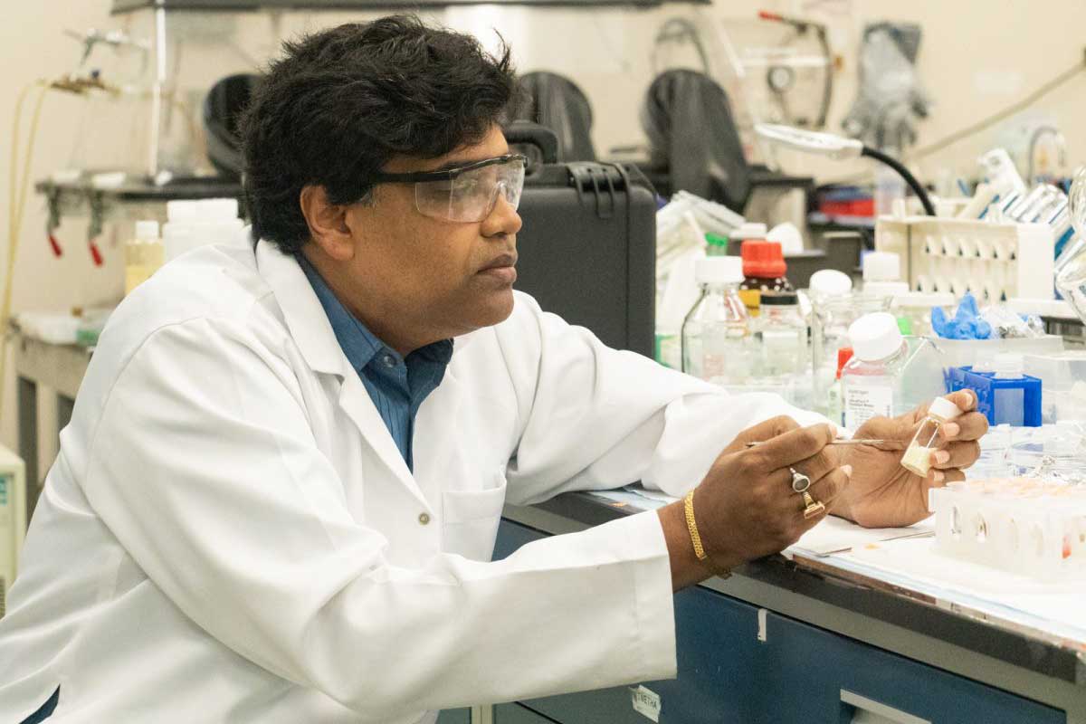 Man in white coat and protective glasses sits at a lab bench
