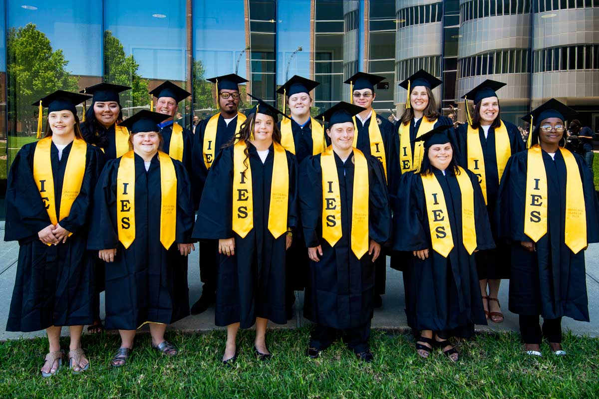 13 students wearing black cap and gowns and gold stolls stand in front of glass building