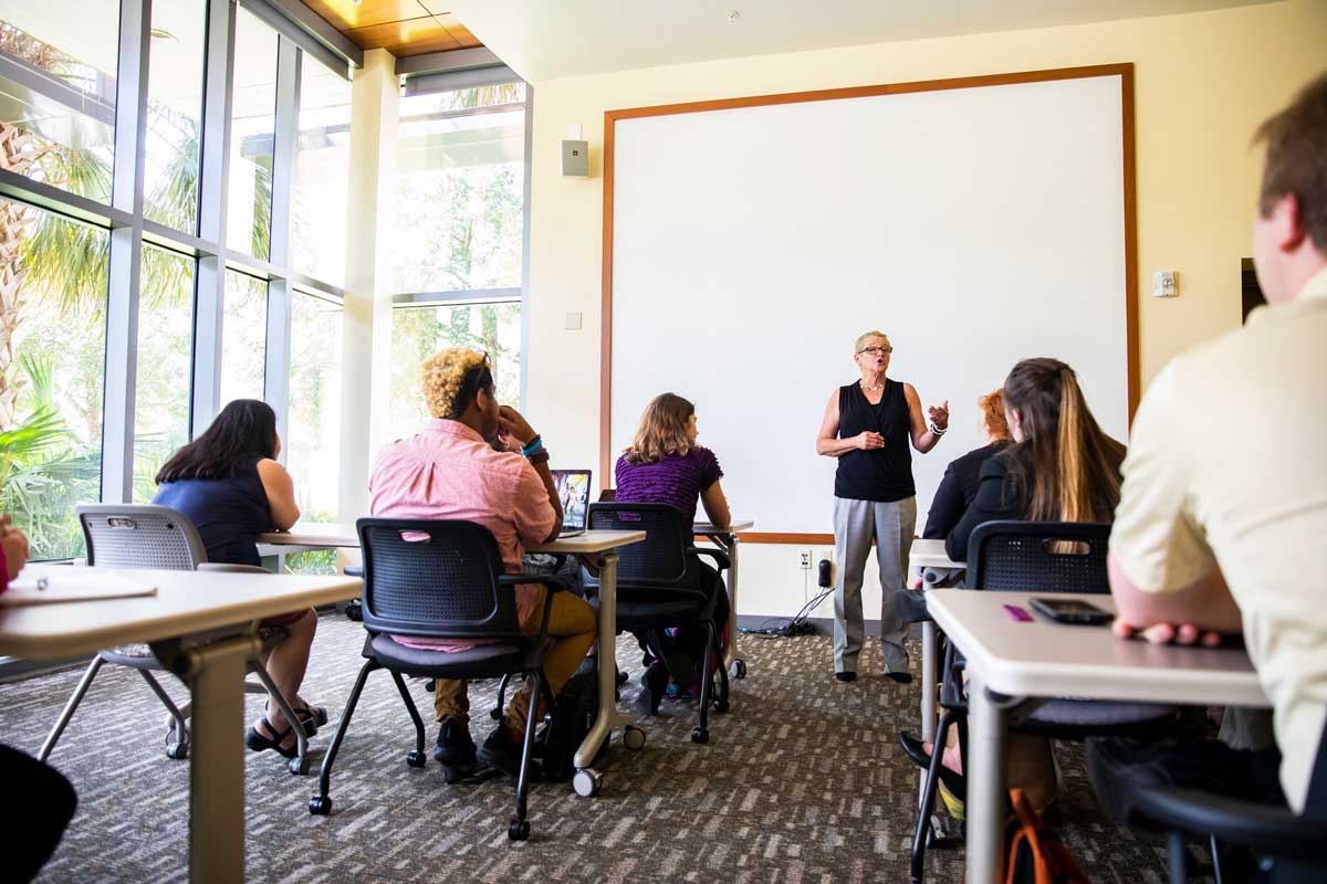 Woman stands at front of classroom in front of blank screen with students sitting at desks in front of her