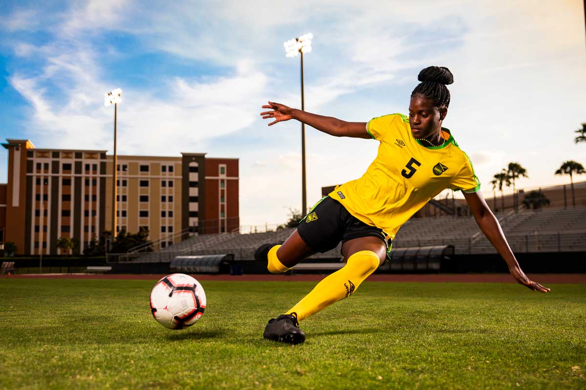 Female soccer player in Jamaica uniform prepares to slide tackle a soccer ball on field 