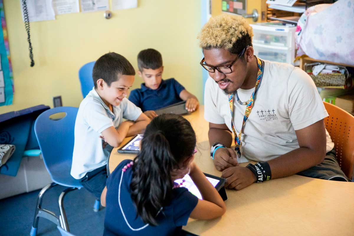 Adult male sits at a table in a classroom with three children holding digital tablets