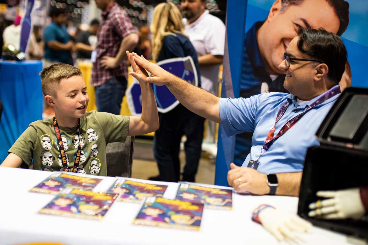 Father and son high five while sitting at a white table with comic books displayed on it.