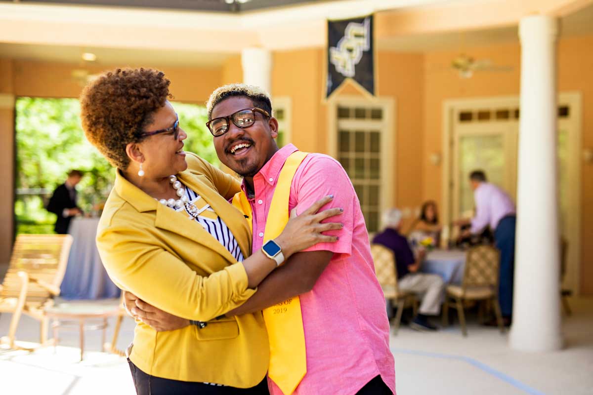 Woman in yellow jacket hugs her son, who is wearing a pink shirt and yellow graduation stoll