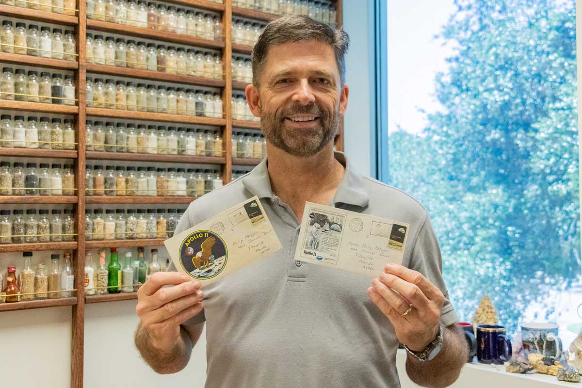 Man with beard wearing gray polo shirt stands in front of dirt samples and holds two post cards in hand