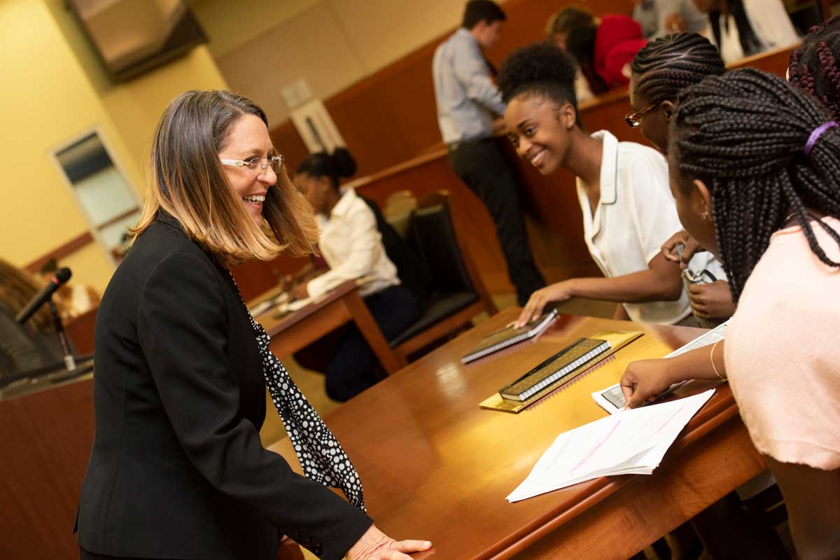 Woman dressed in a business suit addresses teenagers sitting at a wooden table in a courtroom, all smiling