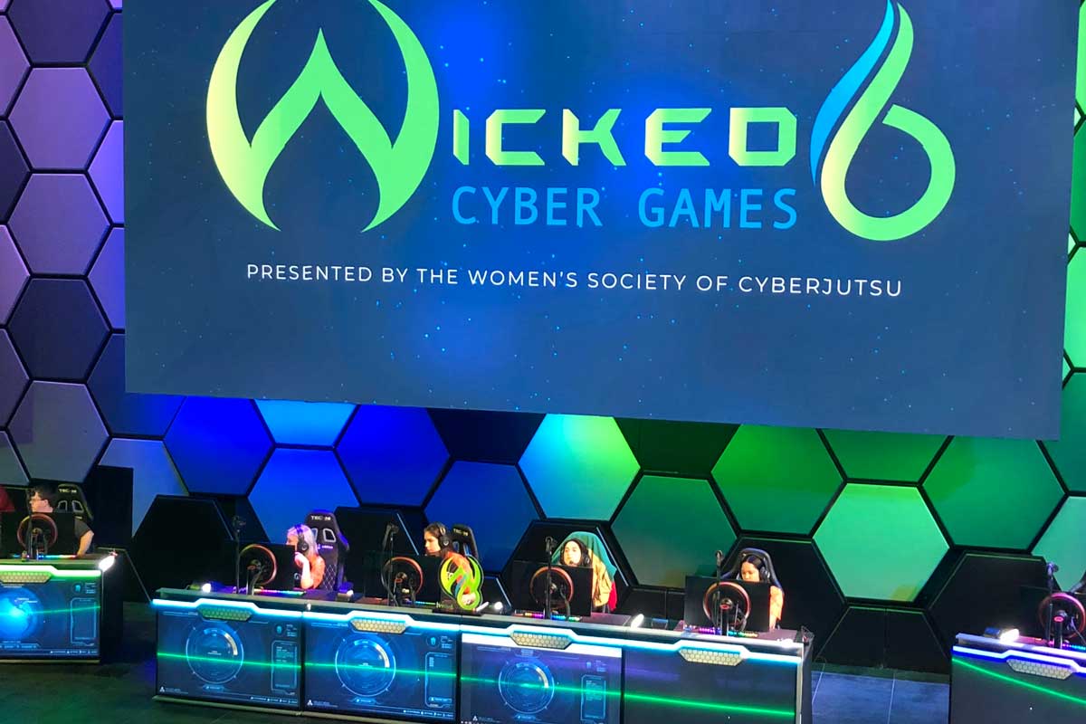Four women sit at a table on blue green stage with headsets on 