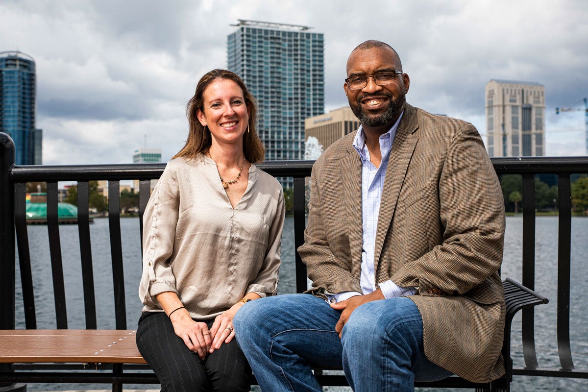 A white woman and a black man who are helping young stroke survivors sit on a bench and talk near Lake Eola