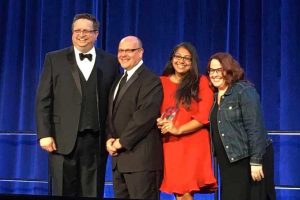 Four individuals pose on stage with glass award in front of blue curtain