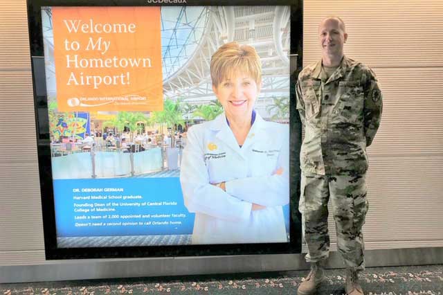 Man in military fatigues stands next to large advertisement in Orlando International Airport