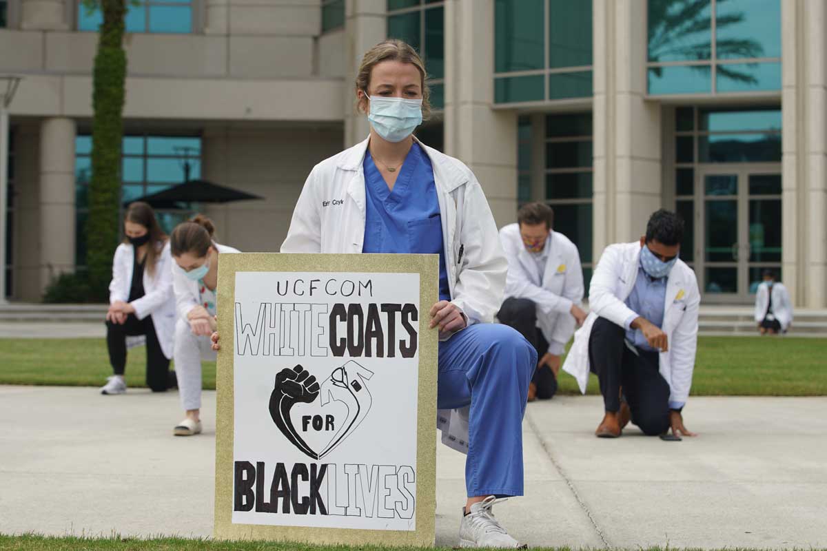 Woman wearing doctor's white coat and face mask holds sign that reads "White Coats for Black Lives"