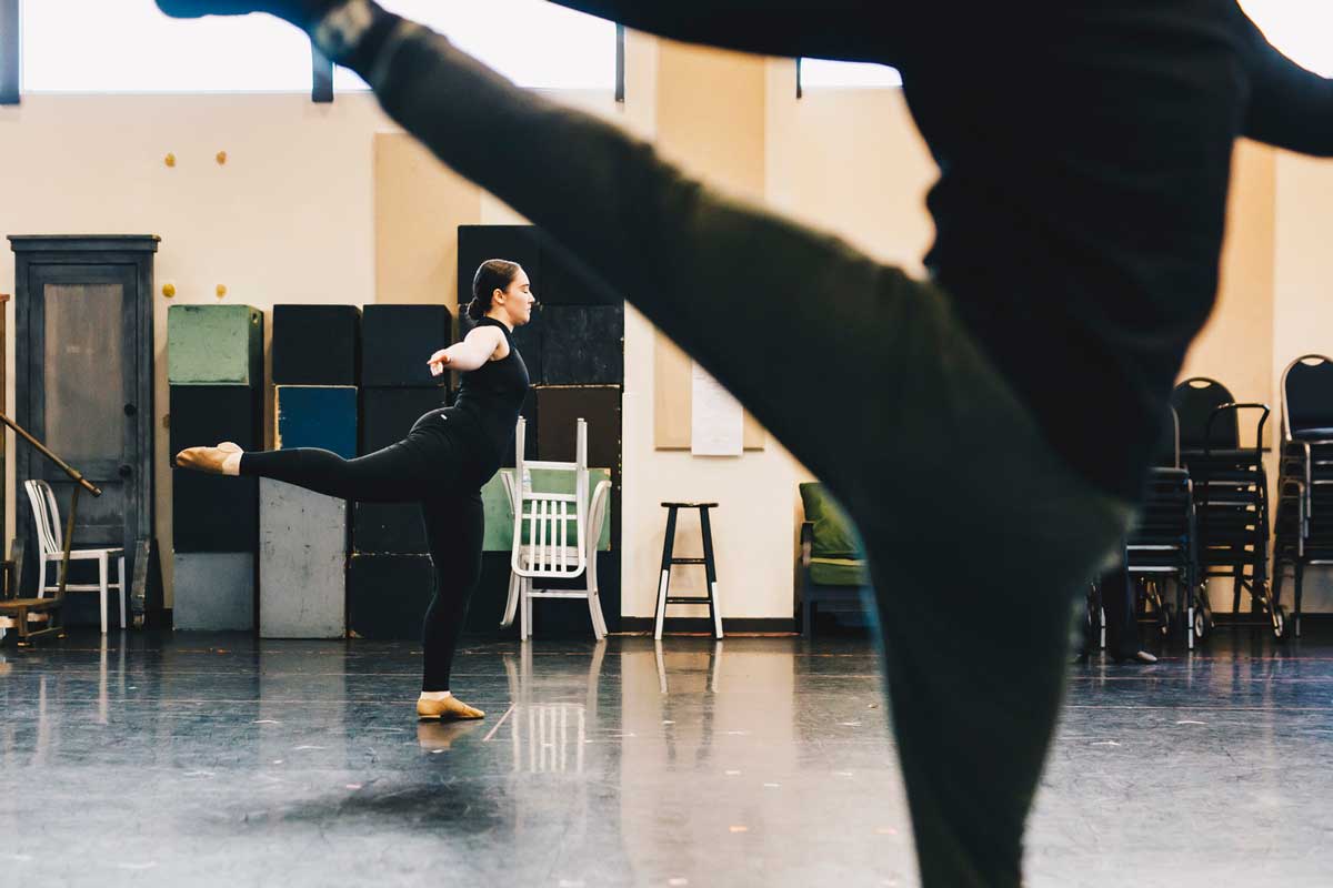 Two dancers dressed in black lift their left legs behind them in dance studio