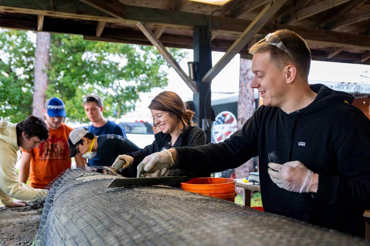 5 students smooth concrete on a canoe under a pavillion