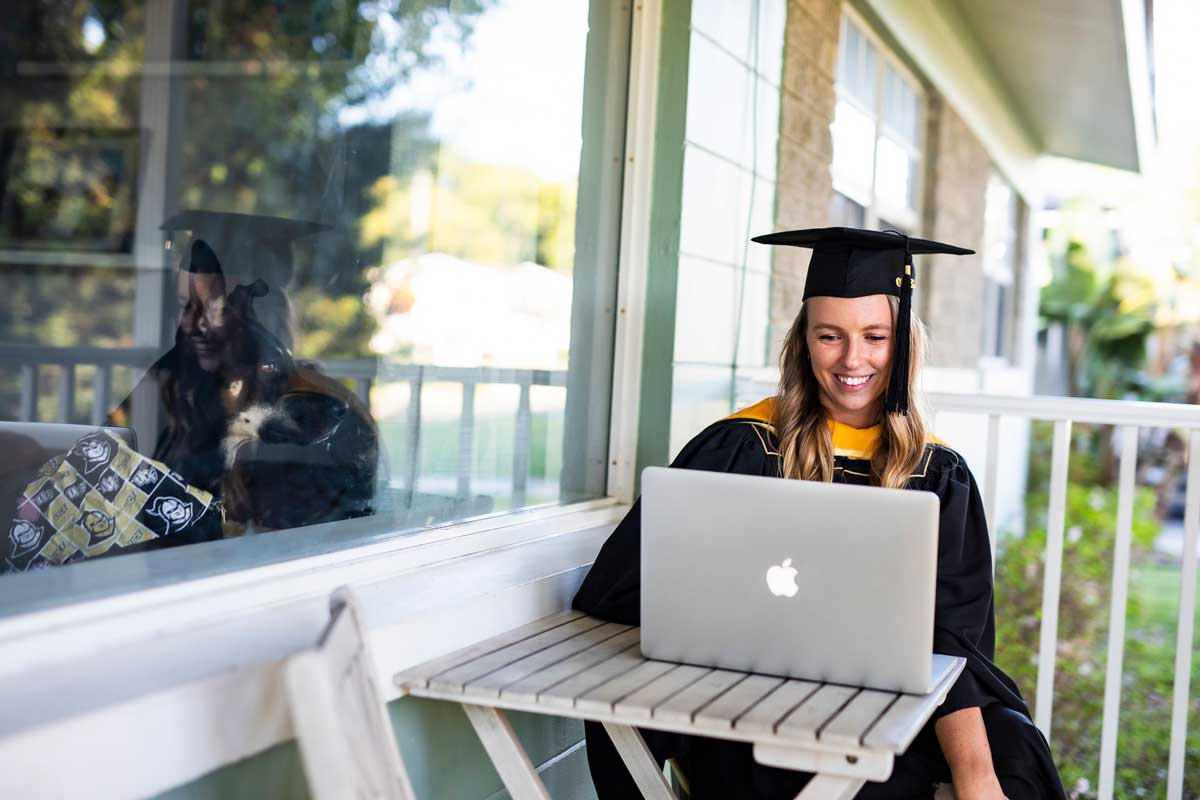 Woman in cap and gown sits on front porch with her Mac laptop as dog inside the home looks at her through a window