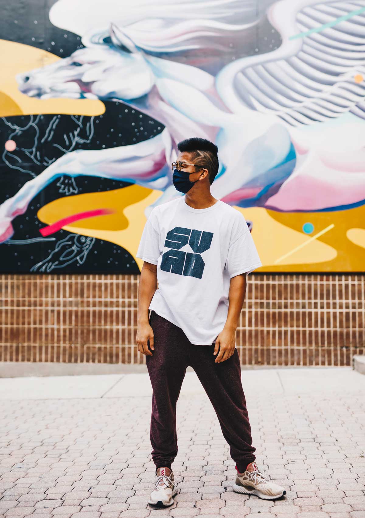 Student wearing black face mask, white shirt and pants stands in front of Pegasus Mural at Student Union